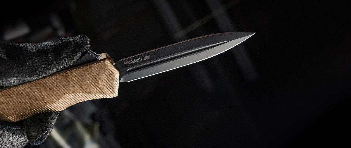 A close-up of a knife with a black blade and a tan textured handle, held by a black-gloved hand against a dark, blurred background. The blade is marked 'MAGNACUT'.